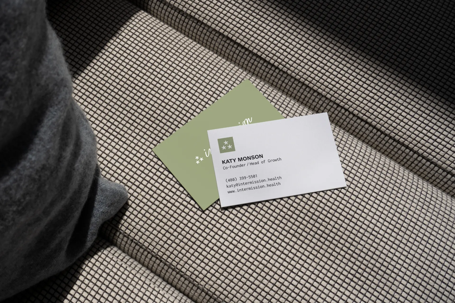 Intermission business cards on a woven fabric surface: a sage green card with the script wordmark and a white card for Katy Monson, Co-Founder / Head of Growth.